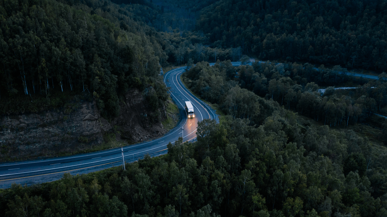 Lastebil kjører på vei i skumring i et landskap av trær.